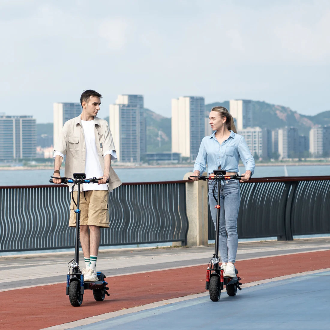 Zwei Personen fahren HITWAY E-Scooter entlang einer Promenade mit Stadtlandschaft im Hintergrund.