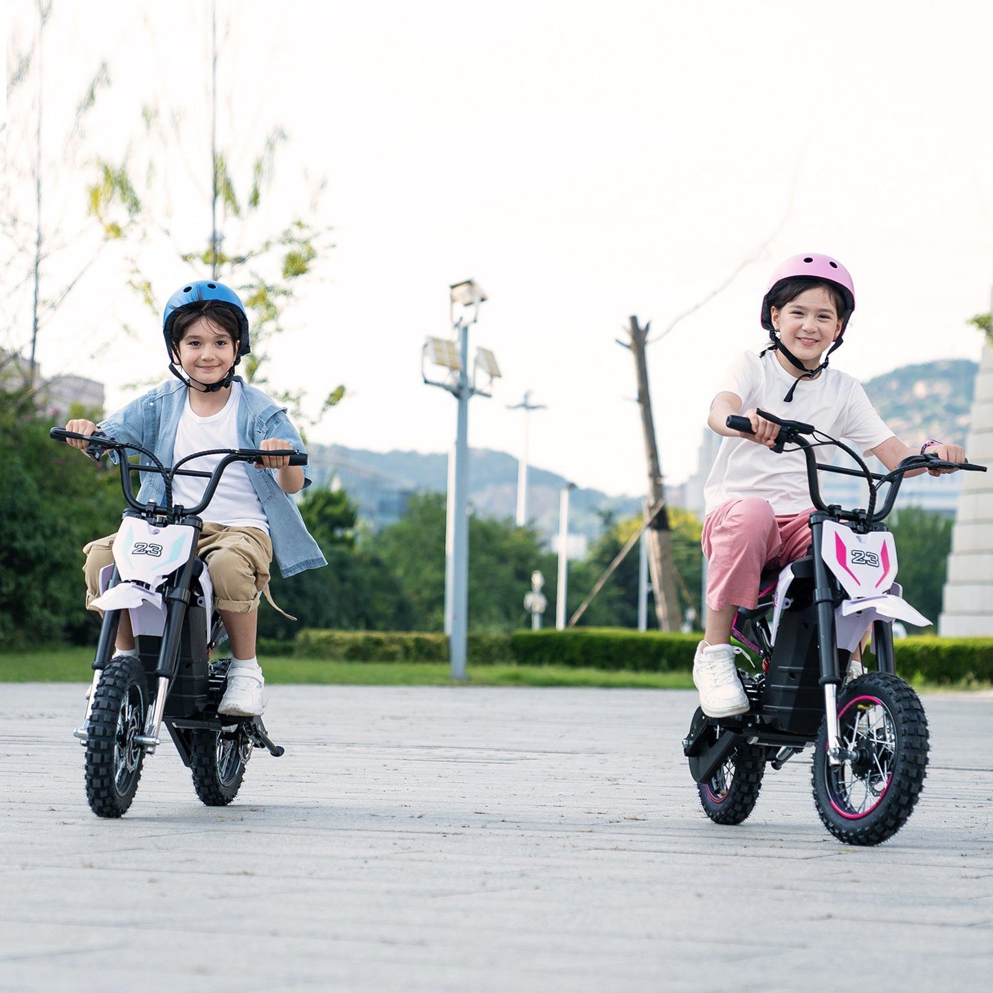 Zwei Kinder fahren das EV65M Electric Kid Motorcycle auf einem Weg, beide mit Helmen ausgestattet, eines in Blau und das andere in Pink, beide lächelnd.